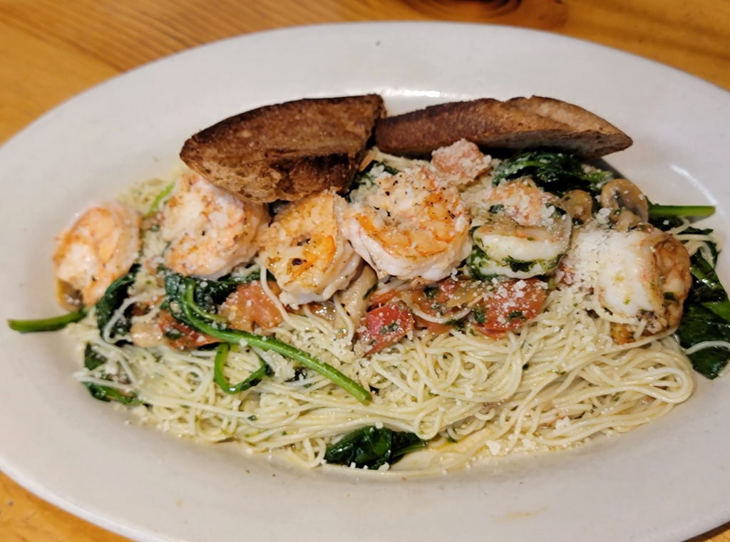 A mound of yellow linguine pasta topped with golden grilled shrimp, and basil pesto sauce sautéed with green spinach leaves, red tomatoes, and brown mushrooms, all served on a white plate. Yum! 