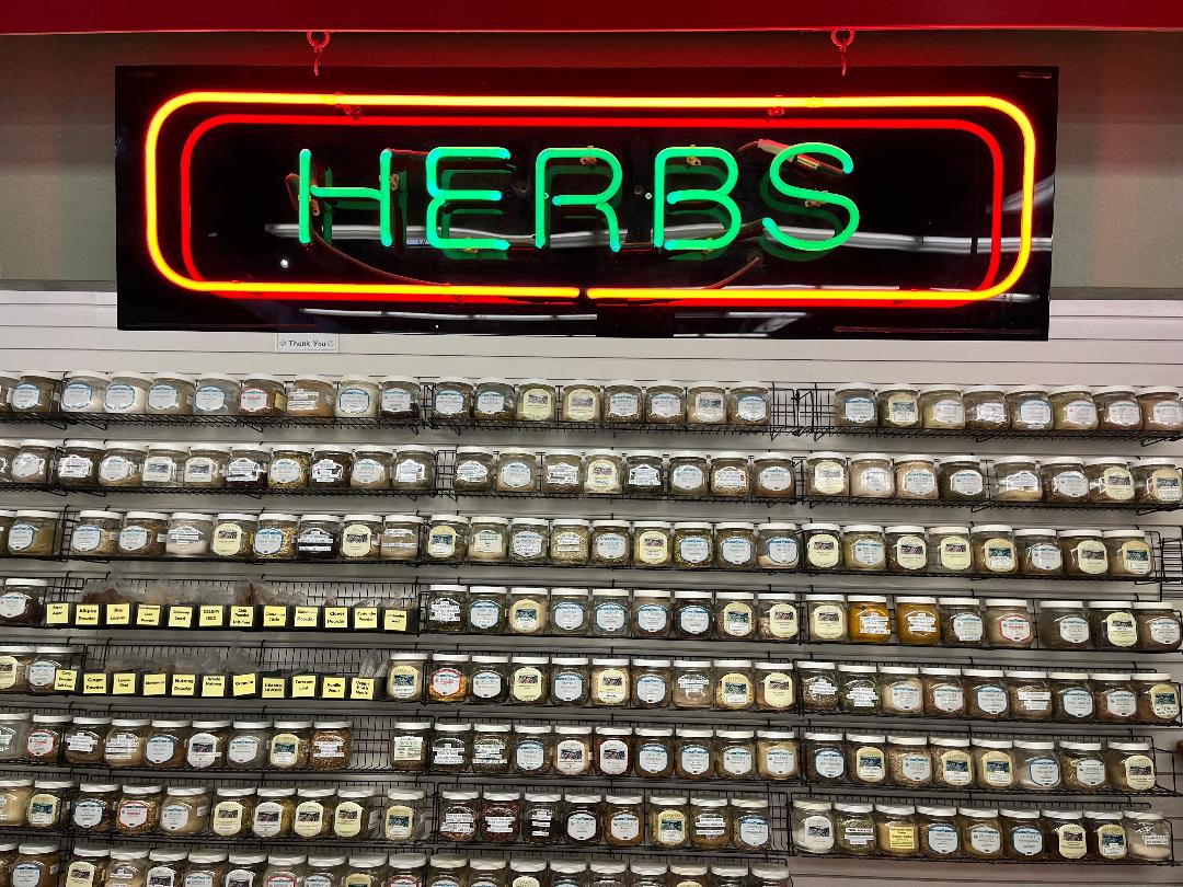 neon sign reading "herbs" in green with a red border, over the top of our herb wall with several rows of jars of bulk herbs and spices.